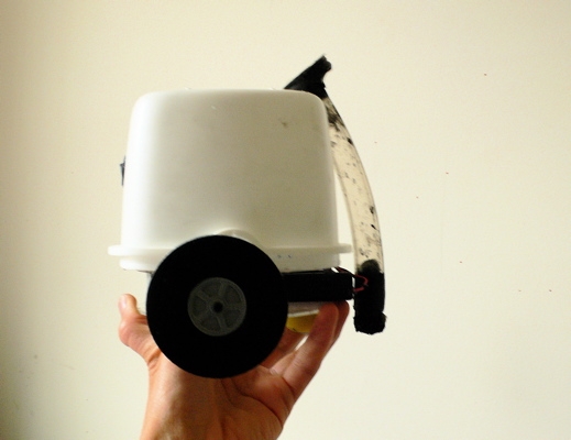 BARB held in one hand: a white inverted Tupperware container with foam wheels, an obstacle-detection sensor on the front, and a brush mount hanging off the back.