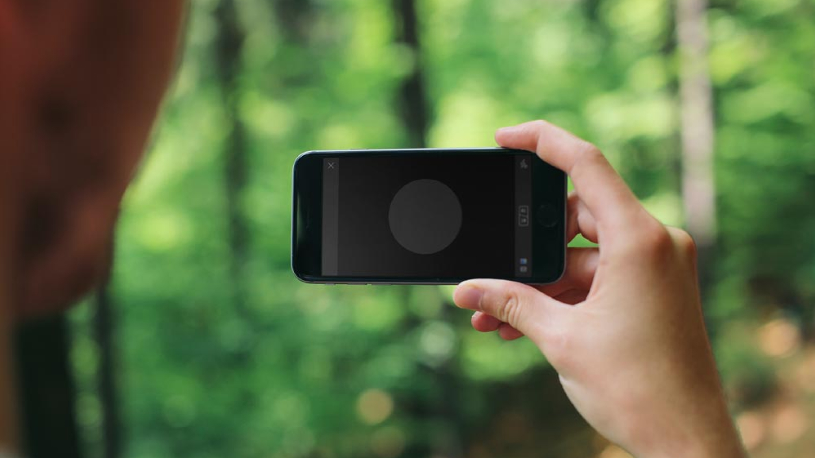 A person holding an iPhone displaying the Shooot app's user interface, with a blurred forest in the background.