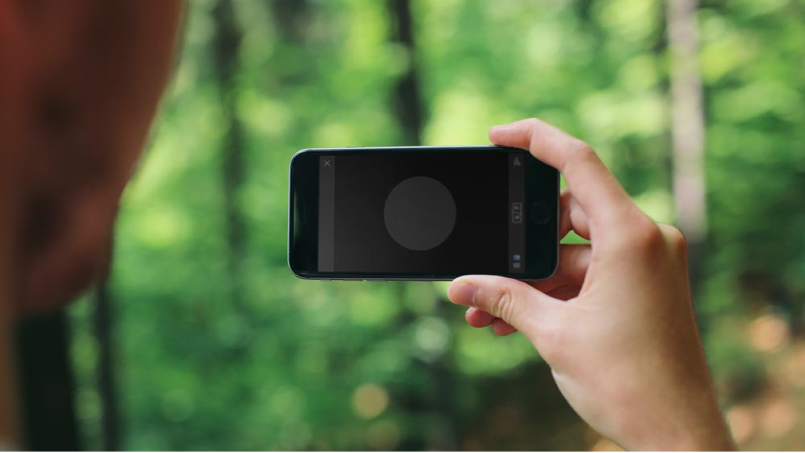A person holding an iPhone displaying the Shooot app's user interface, with a blurred forest in the background.