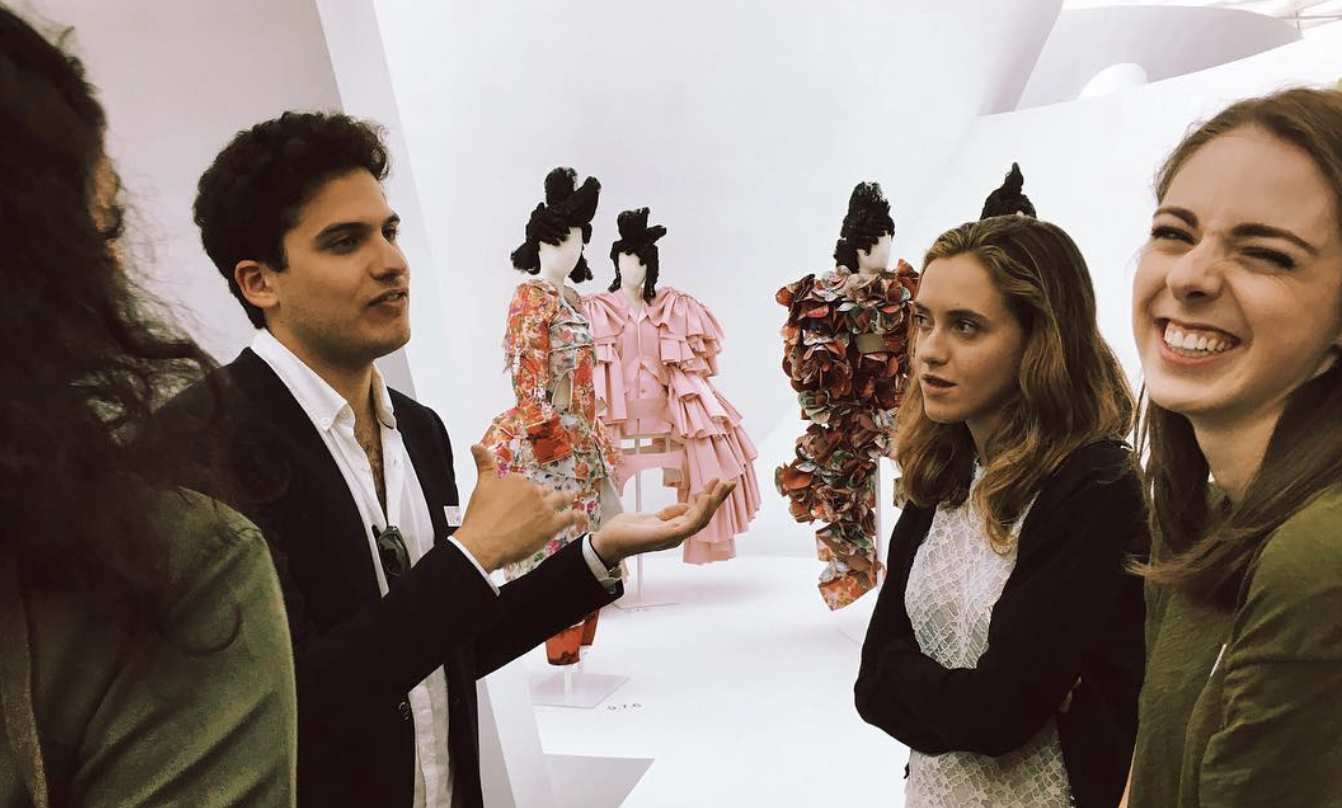 A group of friends laughing and talking inside a fashion exhibit with floral and pink ruffled mannequins in the background.