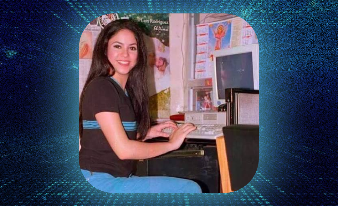 A young woman smiling at the camera while typing at a computer desk, decorated with religious calendars and posters